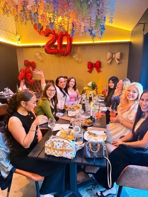 Group of women celebrating a 20th birthday or anniversary at a dinner table with festive decorations and red balloon numerals overhead.