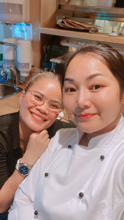 Two women smiling at the camera in a kitchen, one wearing glasses and dark shirt, one in white chef coat