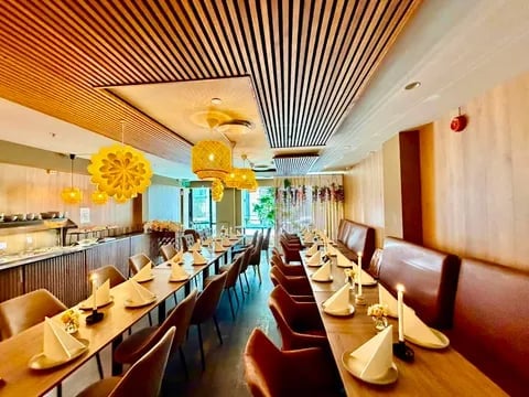 Modern restaurant dining room with long tables, wooden chairs, yellow spherical light fixtures, and decorative striped ceiling.